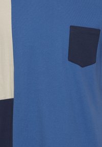 Tissu bleu avec une poche poitrine bleu foncé et des sections partielles crème et marine sur le côté gauche.
