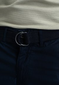 Cintu nero intrecciato con una fibbia metallica circolare, abbinato a una camicia texturizzata beige chiaro. La camicia presenta sottili righe orizzontali.