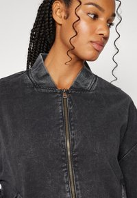 Black denim bomber jacket with a standing collar, zip closure, and ribbed accents. Features a slightly textured surface.