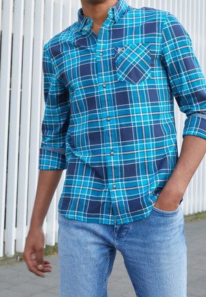 Mężczyzna w niebiesko-zieloną kratę koszulę na guziki z podwiniętymi rękawami i jasnoniebieskie dżinsy, stojący na zewnątrz z jedną ręką w kieszeni.