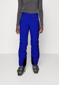 Personne portant un pantalon de ski isolé bleu vif, un haut ajusté à manches longues gris foncé et des bottes de ski noires, se tenant sur un sol blanc.