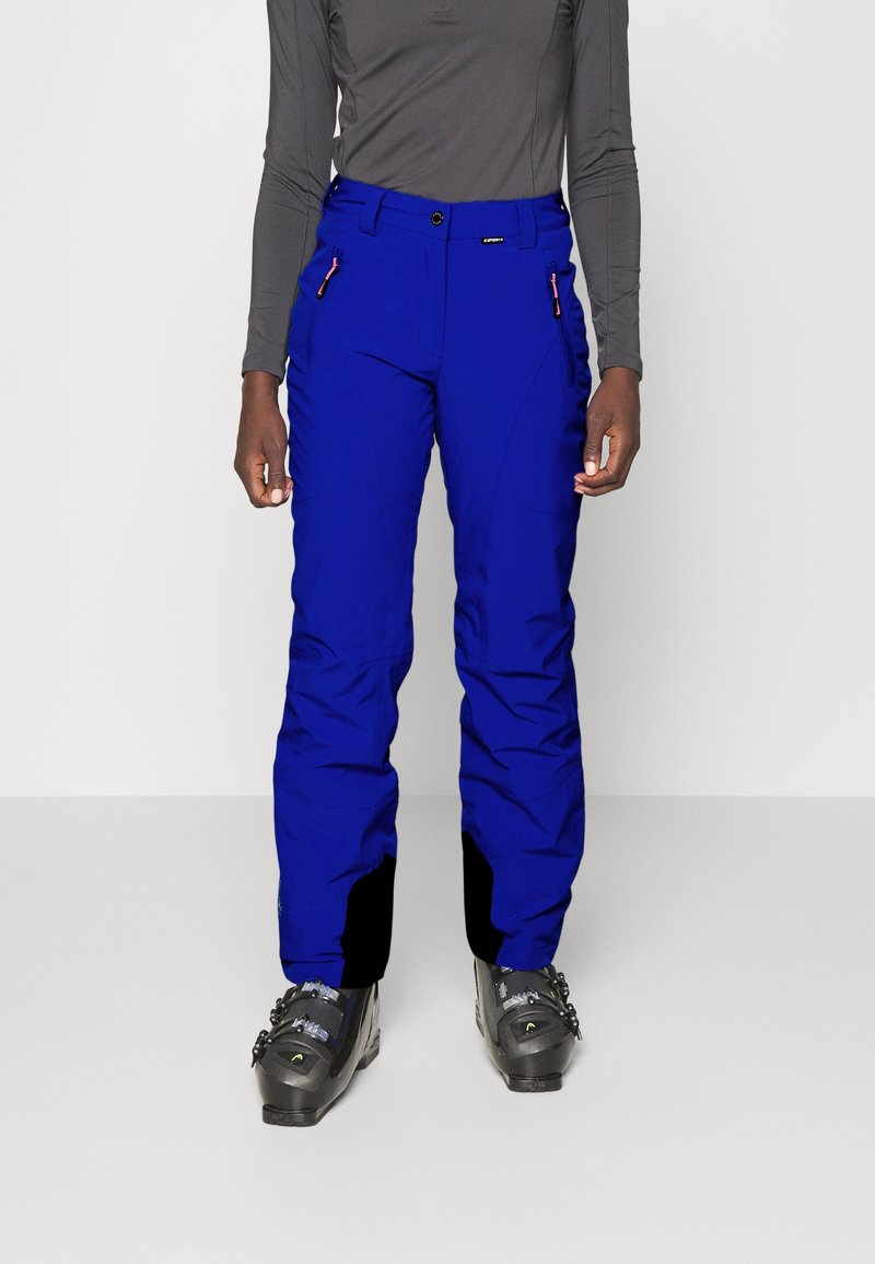 Personne portant un pantalon de ski isolé bleu vif, un haut ajusté à manches longues gris foncé et des bottes de ski noires, se tenant sur un sol blanc.