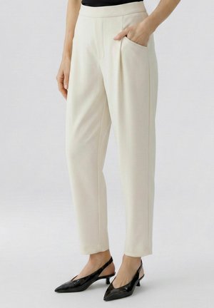 Frau trägt offwhite-farbene, zulaufende Hose mit Seitentaschen und schwarze Slingback-Schuhe mit niedrigem Absatz, stehend vor einfachem Hintergrund.