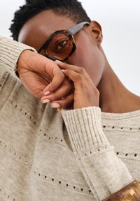 Lunettes de soleil écaille de tortue avec des montures angulaires, associées à un pull léger beige avec des poignets côtelés et des motifs subtils. Teint de peau lisse visible.