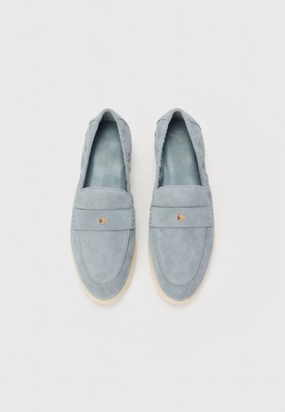 Scarpe slip-on in suede azzurro chiaro con suola piatta e dettaglio in metallo dorato sul davanti. Texture morbida e liscia, con design a punta arrotondata.