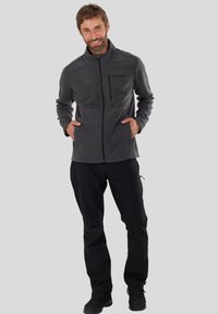 Grijze fleece jas met een hoge kraag, ritszak op de borst en zijzakken. Draagt met zwarte broek en zwarte schoenen.