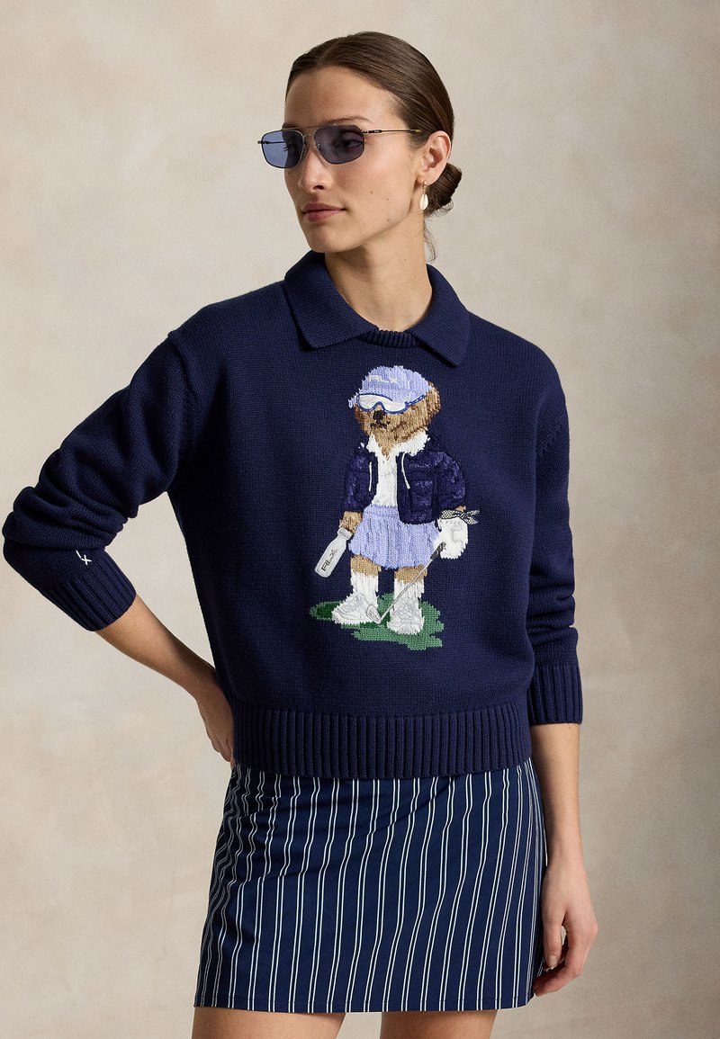 Femme portant des lunettes de soleil bleues, un pull bleu marine avec un motif d'ours jouant au golf, et une jupe rayée verticale bleue et blanche.