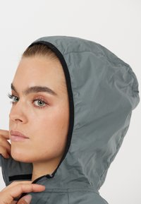 Kevyt harmaa huppari, jossa on musta reunus, varustettu vetoketjulla ja pehmeällä, sileällä pinnalla. Suunniteltu mukavuutta ja helppokäyttöisyyttä silmällä pitäen.