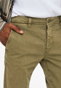 Khaki chinos med en glat tekstur, der har en knaplukning og sidelommer. Stoflet er blødt med subtile folder ved taljen.