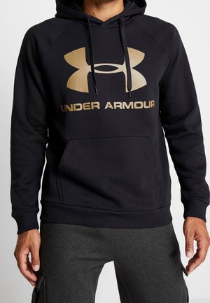 Homme portant un sweat à capuche noir Under Armour avec un logo et un texte dorés, assorti d'un pantalon de jogging gris foncé Puma. Mains détendues le long du corps.