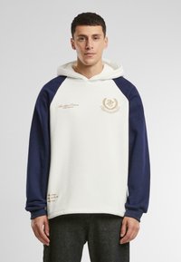 Sudadera con capucha de cuerpo blanco y mangas raglán en color marino. Presenta un logo bordado y acentos de texto en color dorado. Textura de tejido suave.