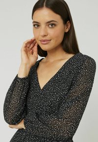 Robe noire avec de longues manches transparentes, dotée d'un col en V et de pois blancs sur l'ensemble. Le tissu texturé ajoute de la dimension.
