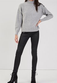 Pull en maille grise avec un col montant et un ourlet côtelé, associé à un pantalon noir ajusté. Bottines noires avec un détail de lacets.