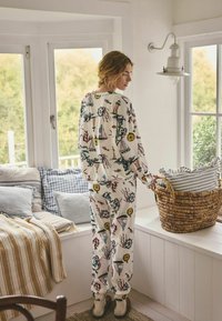 Bílé bavlněné pyžamo s barevnými námořnickými motivy, dlouhými rukávy a kalhotami ve stylu joggerů. Pokoj zahrnuje proutěný koš a polštáře.