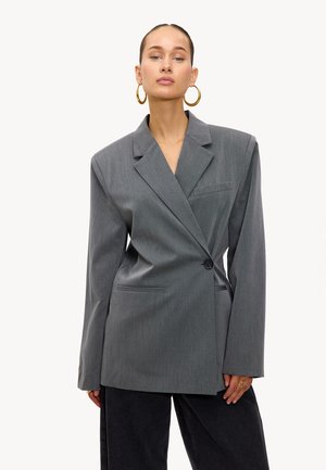 Femme portant un blazer gris surdimensionné avec un seul bouton noir, un pantalon noir, des boucles d'oreilles en or et une bague, se tenant contre un fond blanc.