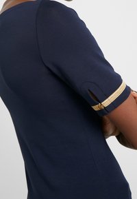 Tmavě modrý krátký rukáv s kulatým výstřihem, s manžetou se zlatými pruhy a bočním výřezem. Vyrobeno z hladké látky.