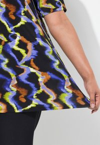 Haut noir et multicolore avec un motif en vagues bleu, vert, orange et jaune. Manches courtes et coupe ample, maintenu par une main au niveau de l'ourlet.