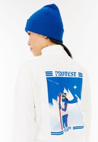 Hvid fleecejakke med høj krave, der har et blåt grafisk design af en skiløber på bagsiden. En blå strikket hue fuldender looket.