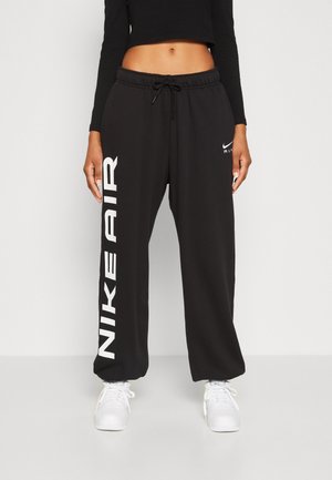 Person trägt schwarze Nike Air Jogginghose mit weißem Logo am Bein und weißen Sneakern, steht vor einem schlichten Hintergrund.