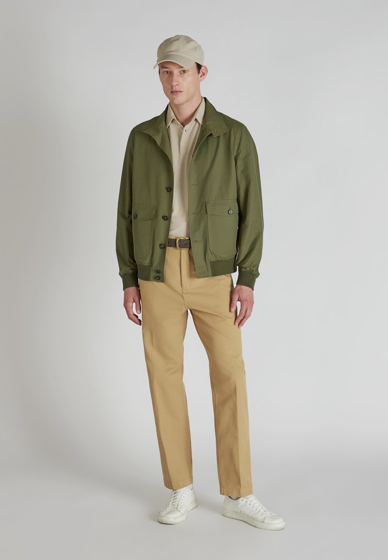 Jasnozielona, lekka kurtka z dwoma przednimi kieszeniami, beżowa koszulka polo, jasnobrązowe spodnie i białe sneakersy, w połączeniu z jasnobeżową czapką.