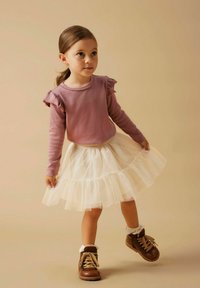 Svijetlopurple dugi rukav majica s naborom, u paru s bijelom tila suknjom i smešnim čizmama sa čipkastim manžetama.