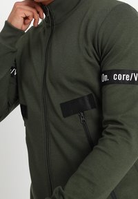 Giacca zip-up verde scuro con colletto alto, caratterizzata da cinturini neri e due tasche con zip sul petto. Tessuto liscio e aderente.