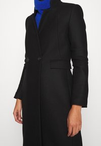 Abrigo negro con un diseño entallado, cuello mao, botones dobles y bolsillos laterales, fabricado en un material texturizado.