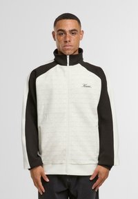 Bela in črna zadrgo jakna z strukturiranim vzorcem, visokim ovratnikom in kontrastnimi rokavi, z vezenim napisom "Kani" v kurent pisavi.