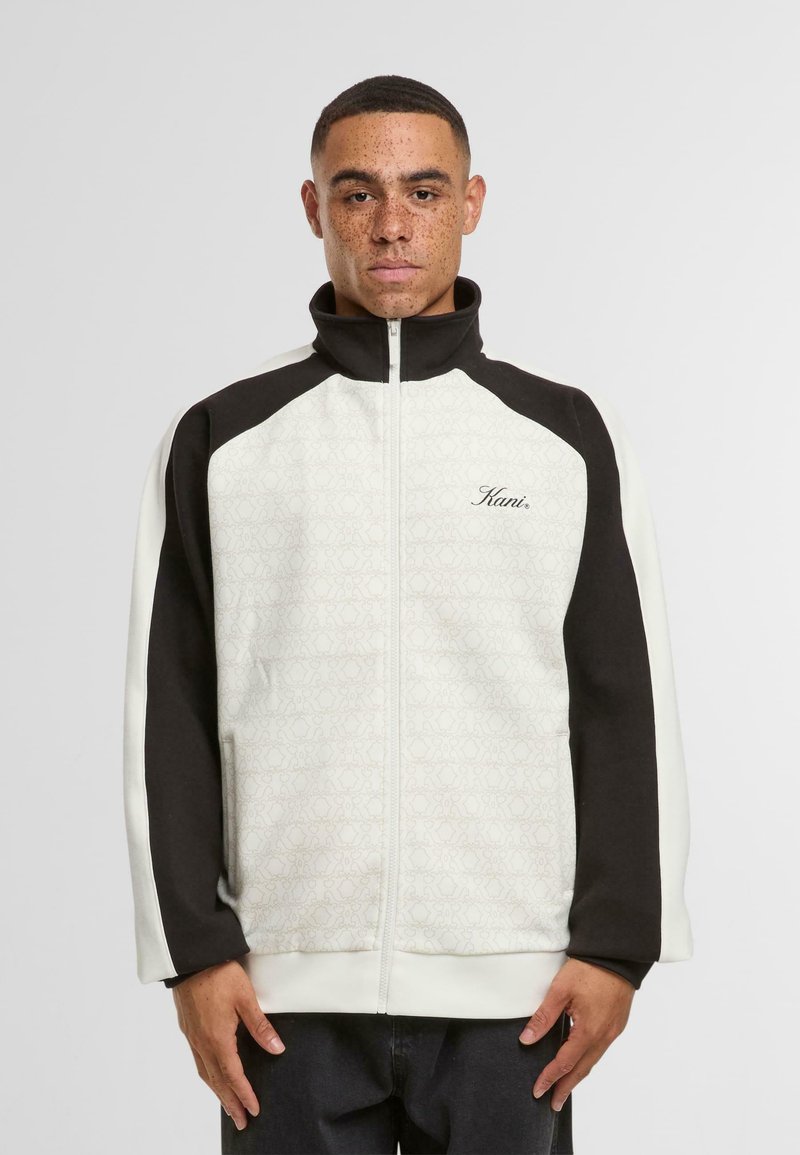 Bela in črna zadrgo jakna z strukturiranim vzorcem, visokim ovratnikom in kontrastnimi rokavi, z vezenim napisom "Kani" v kurent pisavi.