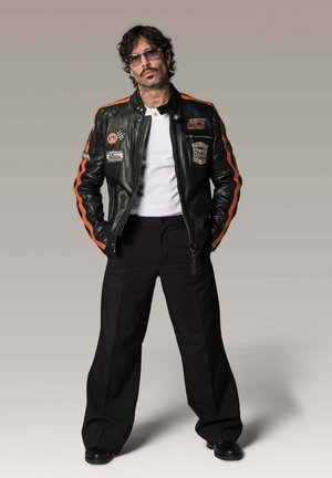 Homme avec des lunettes de soleil portant une veste en cuir noire avec des rayures et des écussons orange, chemise blanche, pantalons larges noirs, debout les mains dans les poches.