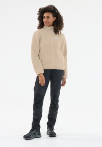 Ljust beige fleece-jacka med hög krage, dragkedja fram och två fickor. Buren med svarta byxor och grå vandringsskor mot en neutral bakgrund.