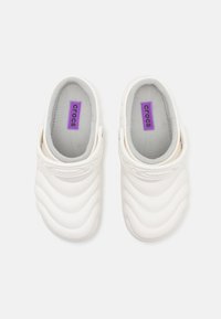 Sabots en caoutchouc blancs avec un design texturé en vagues, intérieur gris et étiquette de marque violette. Style à enfiler avec une sangle à l'arrière pour un bon maintien.