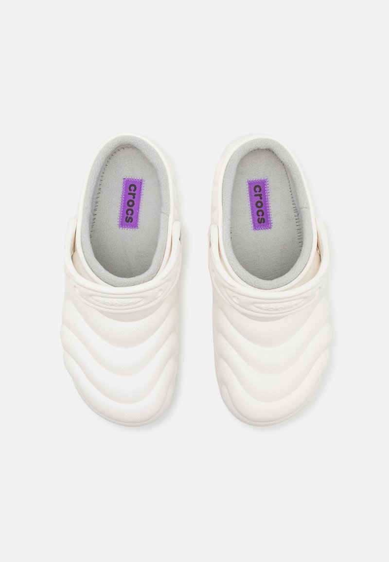 Zuecos de goma blanca con diseño texturizado en forma de olas, interior gris y etiqueta de marca morada. Estilo slip-on con tira trasera para un mejor ajuste.