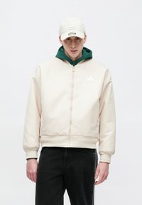 Hele beež värvi pommitusjakk soonikkoes mansettide ja äärega, millel on tõmblukuga sulgemine ning valge kolme triibuga logo. Kandmiseks rohelist kapuutsiga dressipluusi peal.