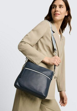 Frau im beigen Anzug trägt eine dunkelblaue Lederschultertasche mit vorderem Reißverschluss und gestreiftem Tragegurt vor weißem Hintergrund.