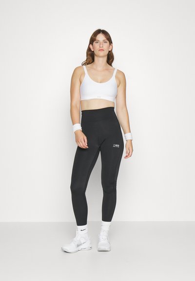 Soutien-gorge de sport blanc associé à un legging noir taille haute avec "2 BADA ACTIVE" imprimé en blanc. Modèle portant des baskets blanches et des bandeaux.
