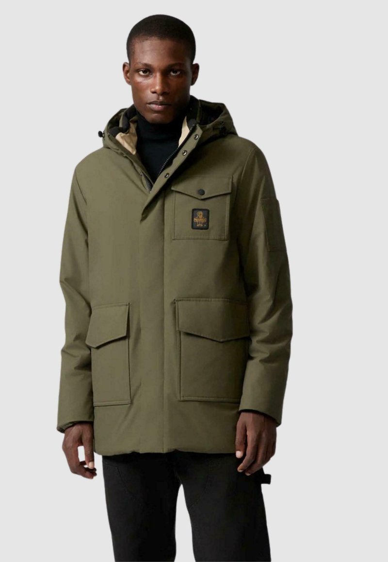 Parka verde oliva con zip frontale, chiusure a pressione, due grandi tasche anteriori e una patch con logo. Presenta un cappuccio e un morbido rivestimento interno.