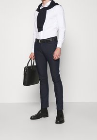 Svart läderväska, vit skjorta med knappar, mörkblå sittande byxor, marinblå tröja hängande över axlarna, svarta skor med en slät yta.