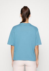 Nainen, jolla on ruskea ponitukka, on pukeutunut löysään siniseen lyhythihaiseen paitaan ja vaaleanpunaiseen housuun, seisoo valkoista seinää vasten, ja hänen kaulassaan ja käsissään näkyy tatuointeja.