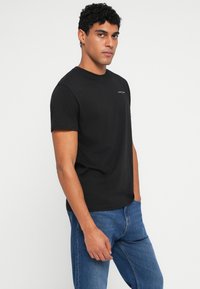 Μαύρο κοντομάνικο μπλουζάκι από βαμβακερό ύφασμα, με στρογγυλή λαιμόκοψη και διακριτική λεπτομέρεια λογότυπου στην αριστερή πλευρά του στήθους. Φορεμένο με μπλε τζιν.