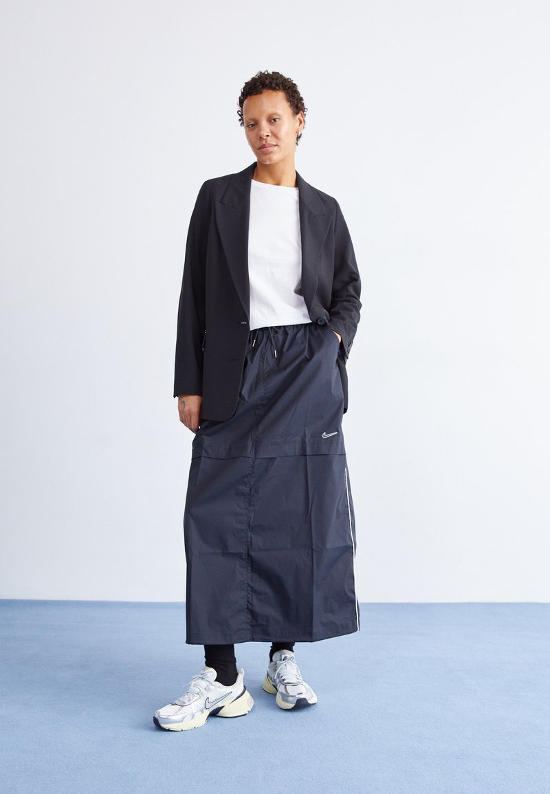 Blazer negro sobre una camiseta blanca, combinado con una falda oscura hasta la rodilla que tiene un cordón y rayas laterales. Zapatillas plateadas con detalles.