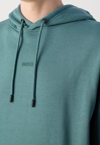 Homme portant un sweat à capuche bleu sarcelle avec cordons et logo "BOSS" en relief sur la poitrine, montré du cou au haut du torse.