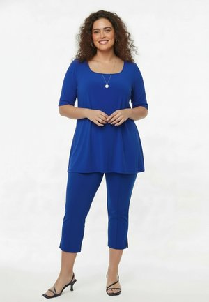 Vrouw met krullend haar, gekleed in een blauwe tuniek top, bijpassende cropped broek, zwarte sandalen met hak en een hangerketting, staand en lachend.