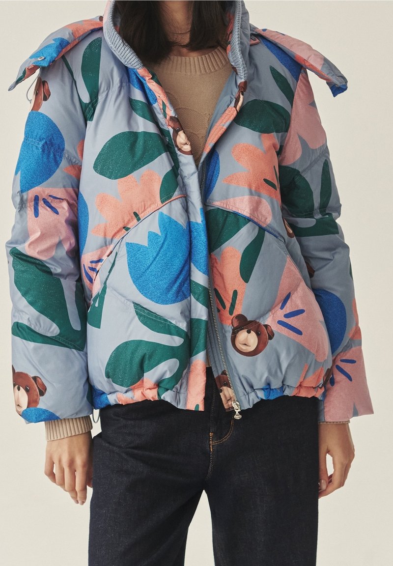 Personne portant une doudoune bleu clair avec de grands motifs floraux colorés et des imprimés ours en peluche, associée à un pull beige et un jean foncé.