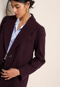 Cappotto in misto lana bordeaux con colletto strutturato, bottoni doppi e revers a scanno, indossato su una camicia azzurra chiara.