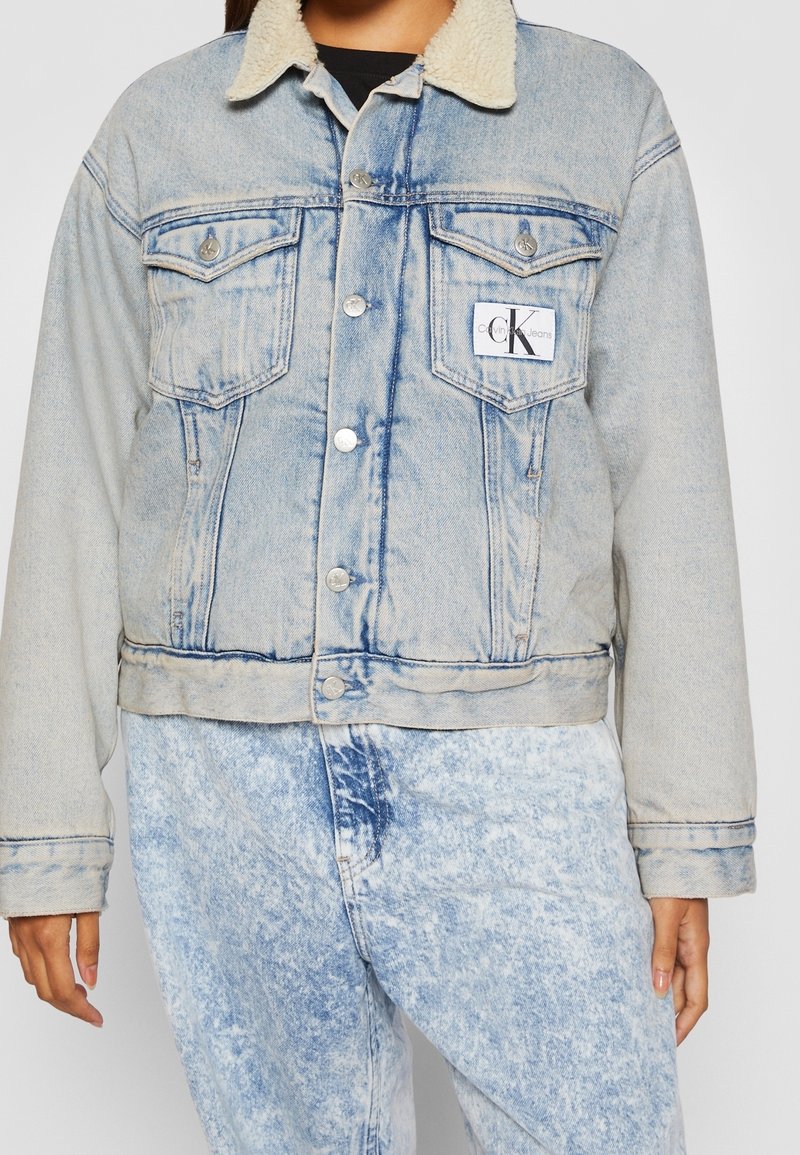 Vaaleansininen farkkutakki, jossa on fleece-kaulus, kaksi rintataskua ja hopeanväriset napit. Yhdistetty vastaavansävyisiin hapotettuihin farkkuihin.