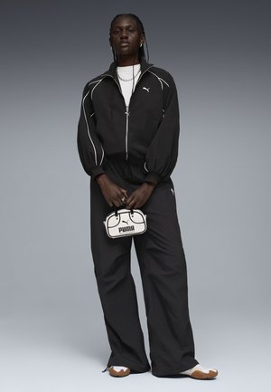 Svart träningsdräkt med vita pipingsdetaljer, zip-up jacka och lösa byxor. Accessoarer inkluderar en liten svart och vit väska med en logotyp.