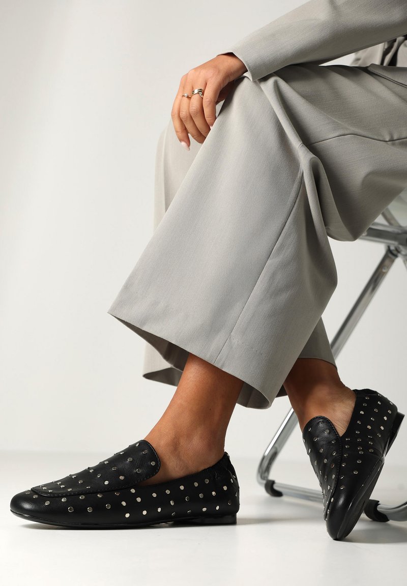 Černé kožené slip-on boty se stříbrnými nýty, kulatou špičkou a plochou podešví. Nosené s světle šedými kalhotami s širokými nohavicemi.