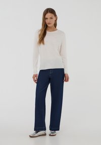Camicia a maniche lunghe di un beige chiaro con sottili decorazioni, abbinata a jeans in denim blu scuro con gamba ampia e sneakers metalliche.