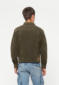 Man met zijn rug naar de camera, gekleed in een olijfgroene denim jas en lichtblauwe cargobroek, tegen een eenvoudige witte achtergrond.